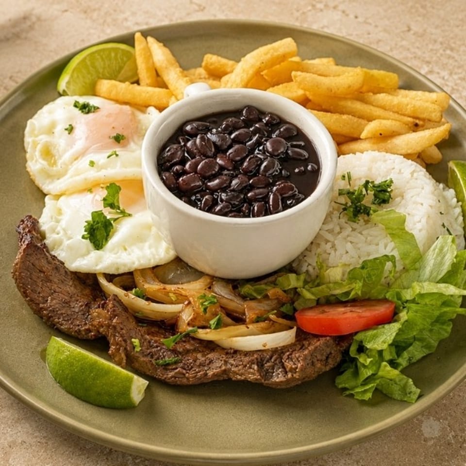 Brazilian-style platter with grilled steak, fried eggs, black beans, white rice, French fries, salad, onions, tomatoes, and lime wedges.