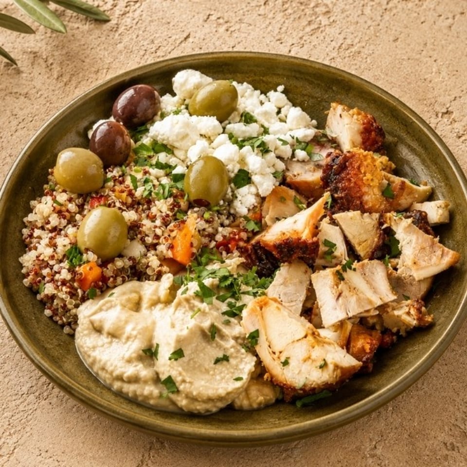 Mediterranean grain bowl with grilled chicken, quinoa, hummus, feta, olives, herbs, and roasted vegetables on a ceramic plate.