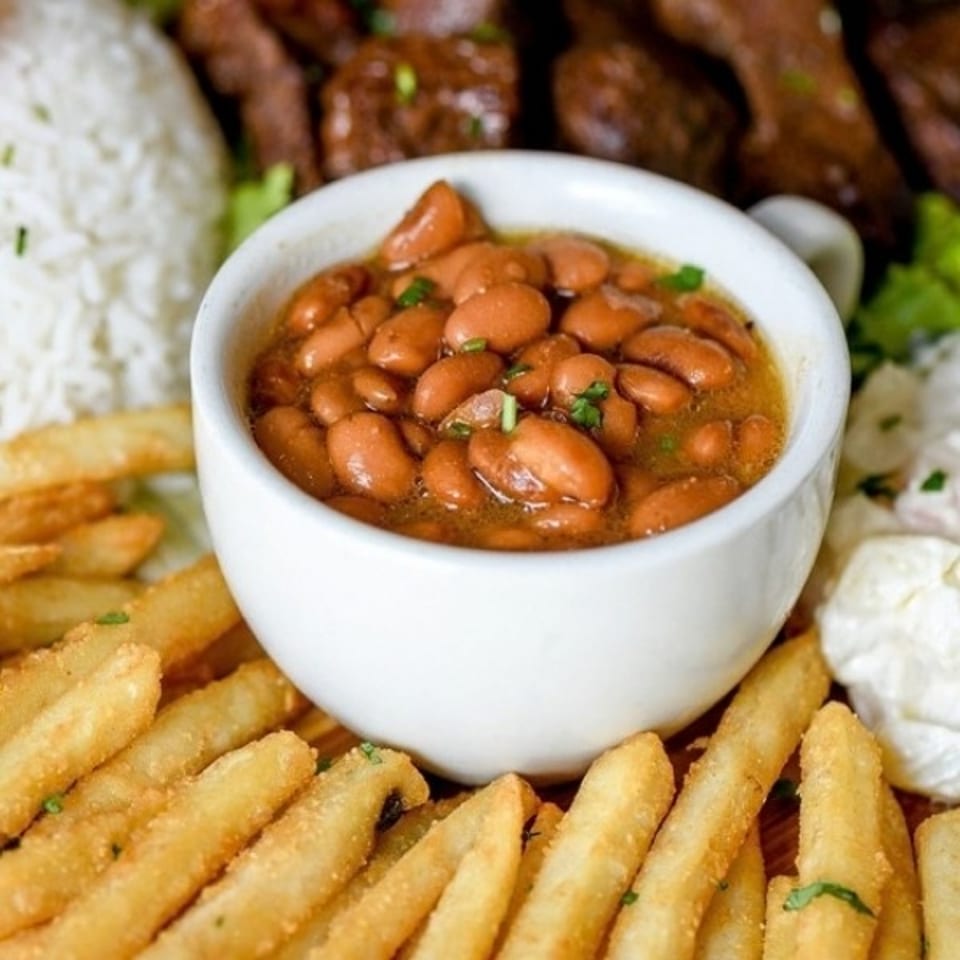 Bowl of seasoned beans surrounded by golden fries, white rice, grilled meat, and creamy coleslaw.